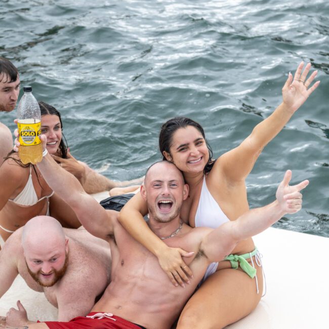 A group of people in swimsuits enjoying a day on the water, smiling and posing for the camera on a boat. One person in the center is holding a cold bottle of Solo. The water sparkles in the background with gentle waves.