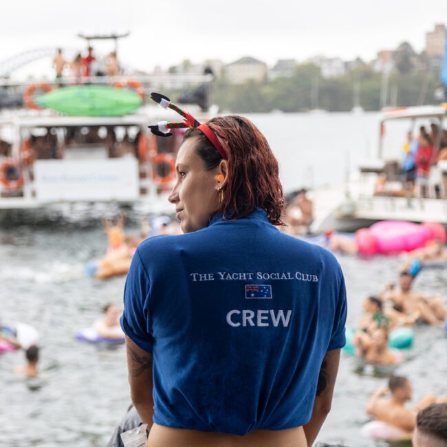 A person in a blue shirt with "The Yacht Social Club CREW" written on the back stands facing a body of water. The background shows people swimming and enjoying themselves on inflatable floats, with boats anchored nearby. Scenic buildings and trees are seen in the distant background.