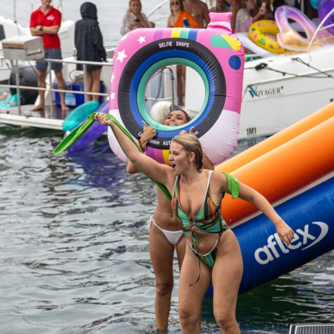 Two women in bikinis enjoy a playful time near a docked boat. One woman has a colorful inflatable ring around her body, while the other energetically gestures with her arm. Other people and inflatables can be seen in the background amidst the lively water, creating a cheerful summer scene.