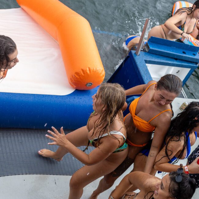 A group of people in swimsuits enjoying a sunny day on a boat. Some are in the water or on inflatable tubes, while others stand on the deck, laughing and socializing. An inflatable slide leads into the sparkling water, creating a fun and exciting atmosphere.