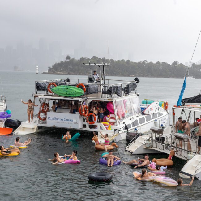 A group of people enjoying a cloudy day on the water with several boats. Many are in beach floats and swim around the boats. A prominent white boat with "Rum Runner Cruises" is in the center, while an impressive cityscape and a large bridge peek through the fog.