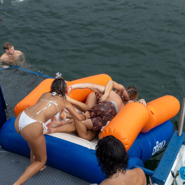 A group of people are enjoying a day on the water. One person is swimming nearby while three others are playfully struggling on a large orange inflatable. A woman in a white bikini joins the fun from the side of a boat, adding to the vibrant scene.