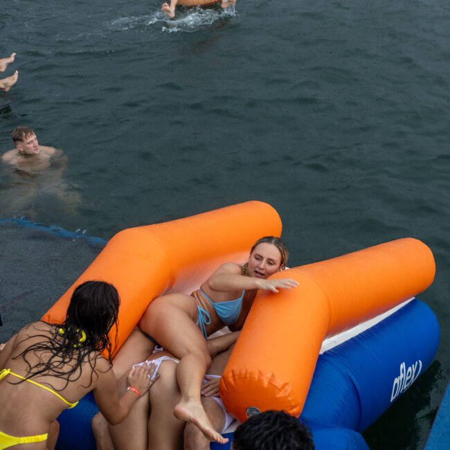 A group of people is enjoying a swim in a large body of water. Some are on inflatable rafts, including an orange and blue one where a couple of individuals are lounging. Others are in the water or standing near the rafts, creating a lively and relaxed atmosphere enhanced by sunlit reflections on the surface.