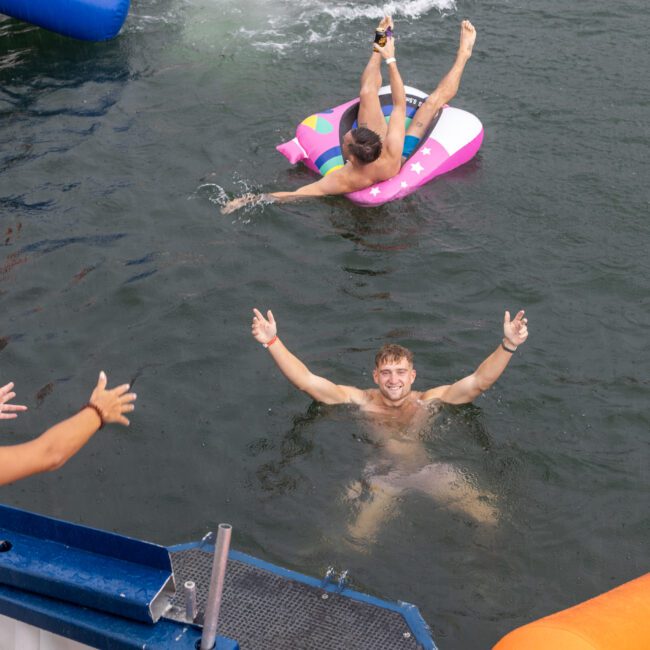 Three friends enjoying a swim, one in the water raising his arms, another on a pink float kicking the water, and a third by the side extending arms toward the swimmer. Brightly colored inflatables, including an adorable reindeer, are floating nearby.