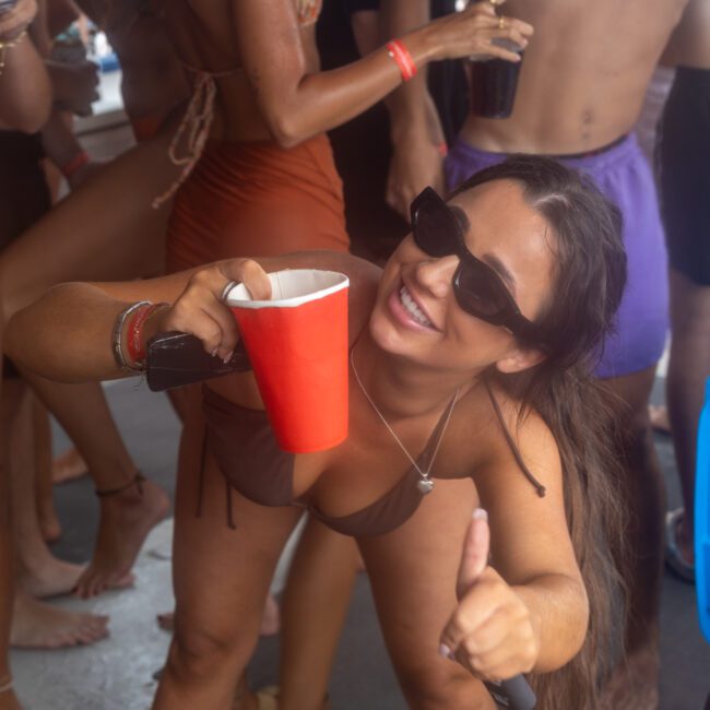 A woman in sunglasses and a swimsuit holds a red cup and gives a thumbs up. She is surrounded by others in swimwear, with a blue cooler visible in the background. The atmosphere appears festive, suggesting a lively pool or beach party full of energy.