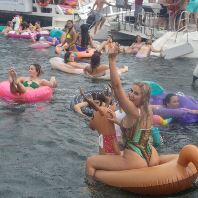 People enjoy a festive time on the water, lounging on colorful inflatable floats shaped like animals and pool toys near boats and yachts. One person waves while sitting on an inflatable unicorn. The scene is lively and filled with joy.