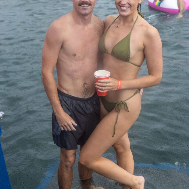 A man in black swim shorts and a woman in a green bikini pose together while standing in shallow water at a beach or lake. The man has a mustache, and the woman smiles while holding a red cup. Inflatable mats and other people are visible in the background.