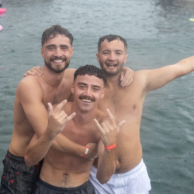 Three smiling men are enjoying a swim in the water, wearing only swim trunks. One man has his arm around another, while the third has his arms outstretched. In the background, a person floats on an inflatable pink ring. The scene appears joyous and festive.