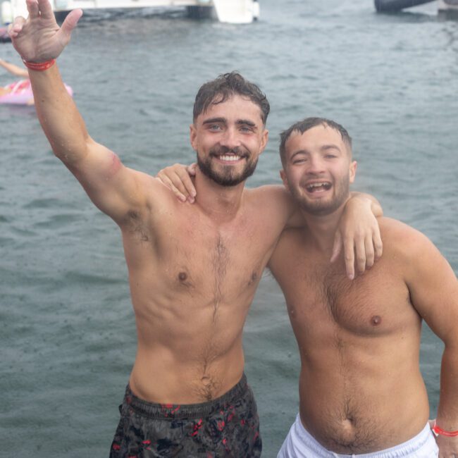 Two shirtless men, smiling and posing arm-in-arm, stand in wavy water near boats. One raises an arm, waving. Both appear joyful and relaxed in a recreational setting. Other people and floating objects are seen in the background.