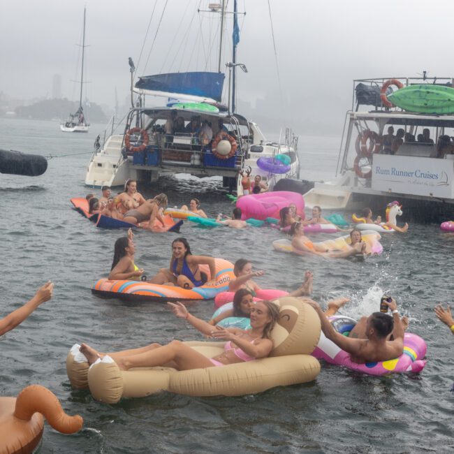 A lively scene of people enjoying a boat party in the rain. They float on colorful inflatable rafts and toys in the water between several anchored boats. Two boats with "Rum Runner Cruises" signs sway gently as revelers soak up the fun beneath a foggy, overcast sky.