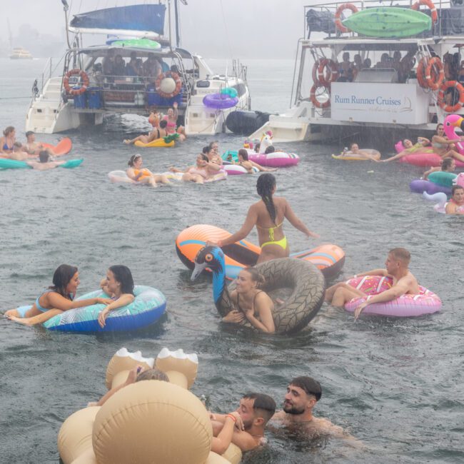 People are swimming and floating on inflatable pool toys in a body of water near several boats. The scene is lively, with individuals enjoying themselves on colorful inflatables like unicorns and flamingos, while a gentle breeze rustles under overcast skies.