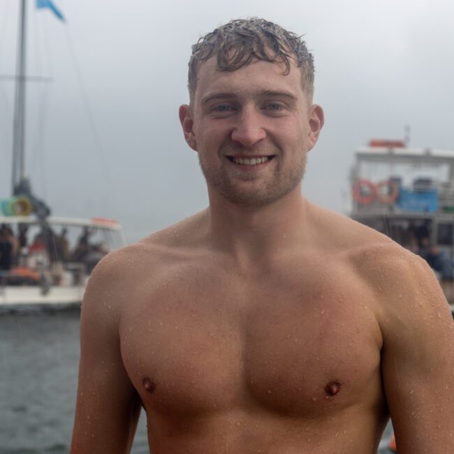 A young man with wet hair smiles while standing shirtless near the water on a gloomy day. In the background, there are blurry boats with people on board and an overcast sky that emphasizes the serene atmosphere.