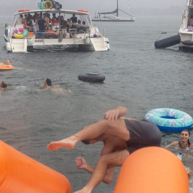 A group of people are swimming and floating on inflatable tubes in a body of water, with two boats nearby. In the foreground, two individuals are mid-air, seemingly jumping or falling off an orange inflatable structure. The atmosphere is lively and energetic as music plays from one of the boats.