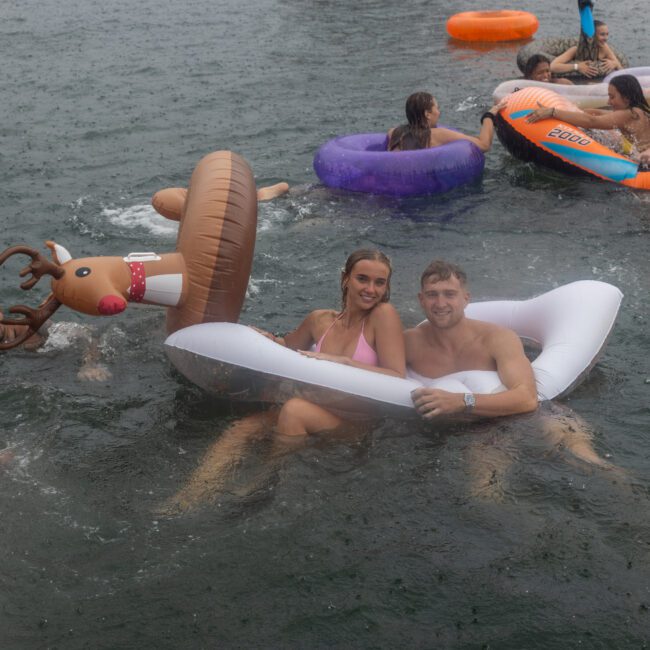 A group of people revels in the water on inflatable floats at a beach party. In the foreground, a couple smiles while sitting together on a white float shaped like an inflatable chair. Others enjoy themselves on colorful floats in the background amidst laughter and waves.