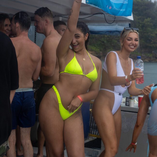 A group of people enjoy a boat party. In the foreground, two women in bikinis dance and smile cheerfully. One woman in a neon green bikini raises her arm, while the other in a white bikini holds a drink. The lively atmosphere is enhanced by music playing as people laugh and mingle.