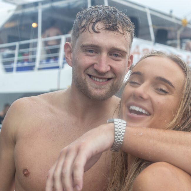 A close-up of two smiling people, a man and a woman, who are wet, possibly from swimming. The man has his arm around the woman. They are aboard a yacht with other people in the background. The atmosphere suggests a fun, relaxed party on the water.