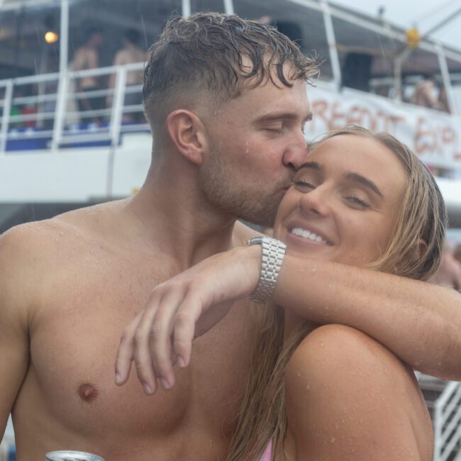 A shirtless man kisses a smiling woman on the cheek while they embrace in front of a crowded boat, both wet from swimming. The vibrant scene is filled with laughter and joy, with people enjoying themselves in the background.