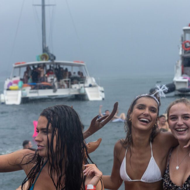 A group of young women in swimsuits enjoying a rainy outdoor party on the water. One of the women is wearing a headband with a small crown, adding to the festive atmosphere. In the background, boats filled with more people celebrating create a lively scene.