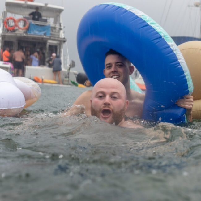 Two men are in the water holding inflatable rings. The man in the foreground has a blue inflatable ring around his neck, swimming towards the camera with his mouth open. In the background, a boat and other people are enjoying themselves amidst a lively atmosphere, creating memorable summer fun.