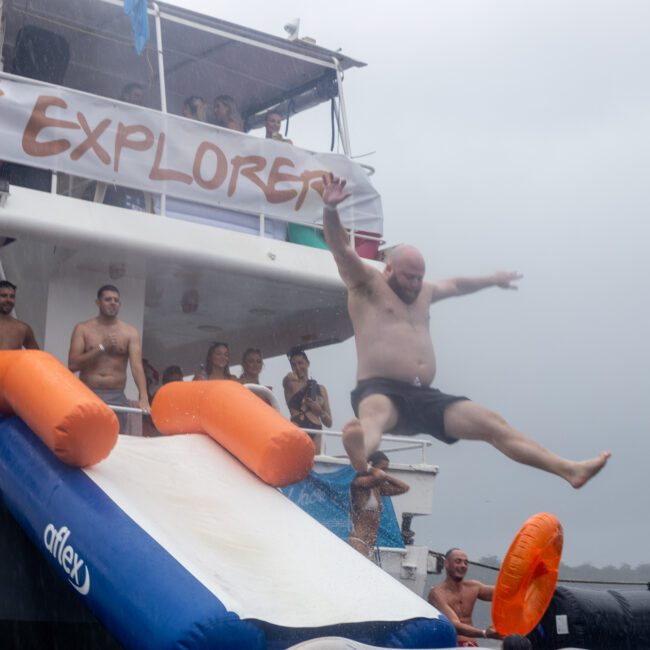 A man is mid-air after sliding off an inflatable slide attached to a boat named "Foot Explorer." He is surrounded by people in swimsuits, some watching and others preparing to slide. The Yacht Social Club Sydney Boat Hire provides a party atmosphere despite the gray and overcast sky.