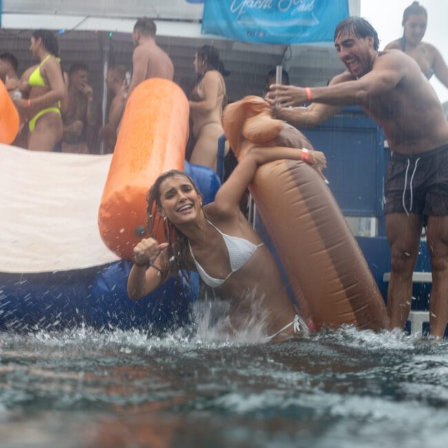 People enjoy a water activity on a floating platform, slipping into the water. A woman in a white bikini slides down an inflatable slide, holding a large inflatable swan, while a man behind her playfully pushes. Others watch and wait their turn with anticipation, eager for their chance at the fun.