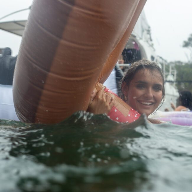 A woman smiles while partially submerged in water, holding onto a large inflatable unicorn. In the background, more people are seen enjoying water activities near a boat and a tree-lined shore. The scene appears festive and joyful.