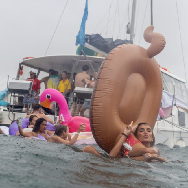 A group of friends enjoy a lively party in the water near a boat, floating on various inflatable toys, including a pink flamingo and a giant brown-ring float. The sky is overcast, and the boat in the background has several people on board, some holding drinks and taking photos.