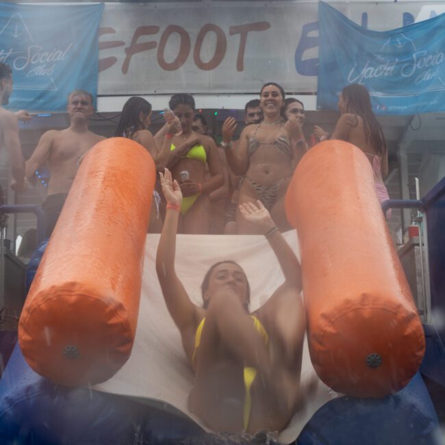 A person in a yellow swimsuit slides down an inflatable orange and white water slide. In the background, several people in vibrant swimwear stand on a platform under a banner that reads "Yacht Social Club." The atmosphere brims with lively, fun energy.