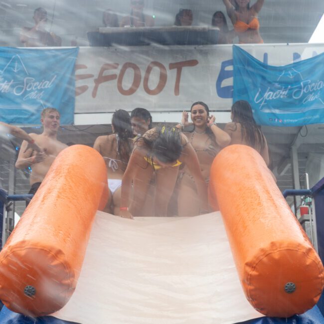 A group of partygoers in swimsuits enjoy a waterslide at the Yacht Social Club event. Two people stand on the platform at the top of the slide, while others gather at its entrance. Bright banners and a light spray of water create a festive atmosphere.