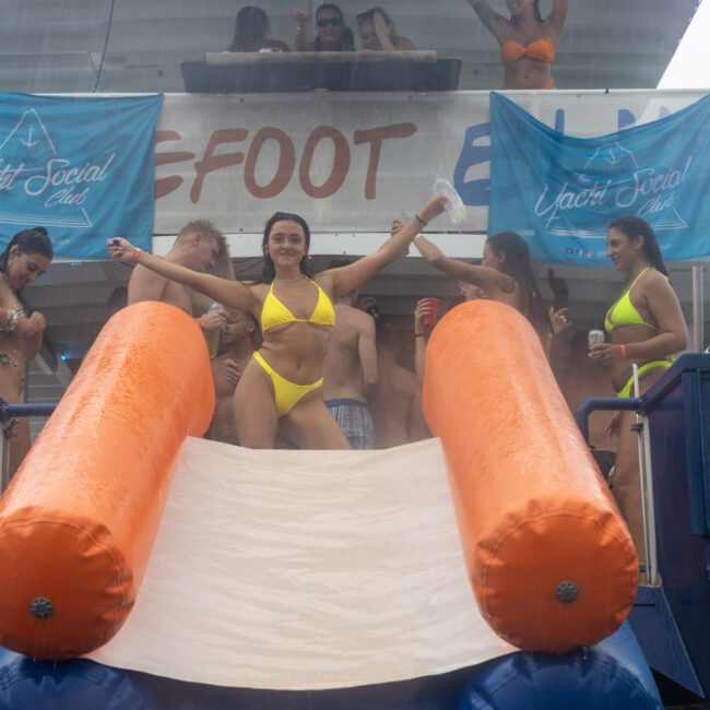 A woman in a yellow bikini poses with arms outstretched at the top of an inflatable slide on a boat, surrounded by people in swimsuits. Blue banners with "The Yacht Social Club" logo are prominently displayed. Other guests stand behind her, watching the lively scene unfold.