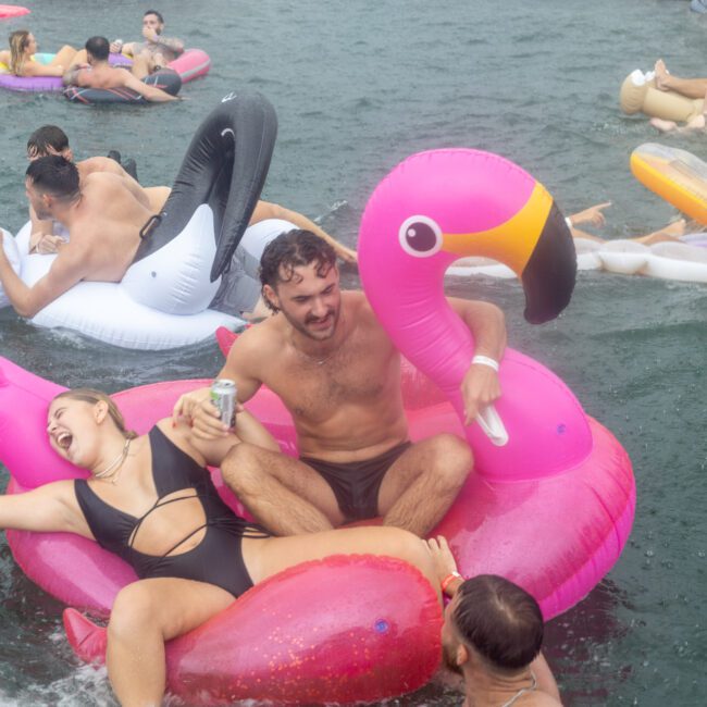 Several people are enjoying a day in the water on various inflatable pool floats, including a colorful unicorn. They are laughing, holding beverages, and having fun in a lively, social atmosphere. The background shows others on similar floats.