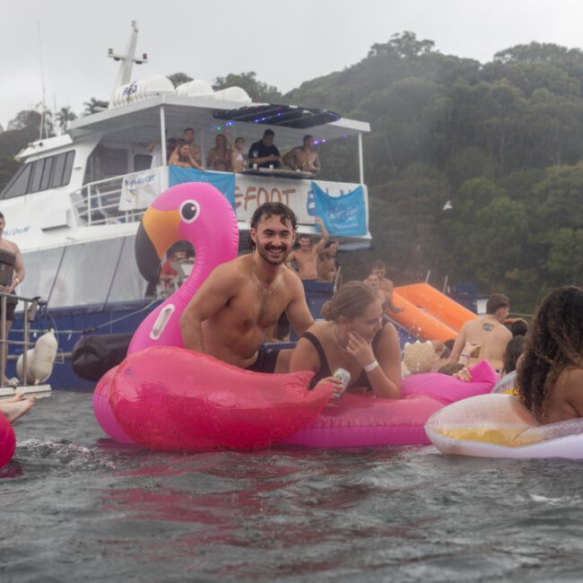 A group of people enjoying a fun day in the water with inflatable floats, including a large pink flamingo float. In the background, cruises with more people are anchored near a forested shore. The sky appears overcast, suggesting light rain.