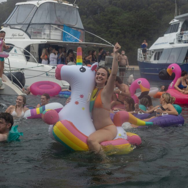 People enjoy a lively gathering at a yacht party, with some swimming and others lounging on colorful inflatable floats shaped like unicorns and flamingos. Sailboats create a picturesque background, and everyone appears to be having a fun time in the water.