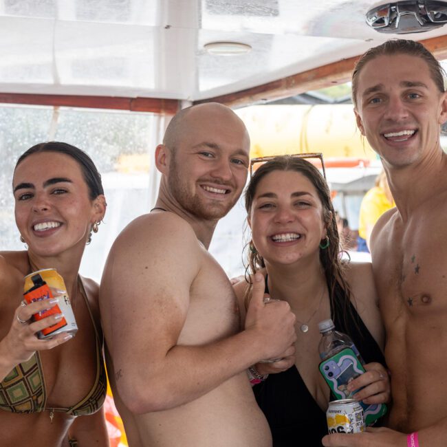 Four people in swimwear are posing and smiling on a boat with drinks in hand. Two women are on the left and right ends, while two men are positioned between them. The background shows a beautiful view beyond the boat's interior.