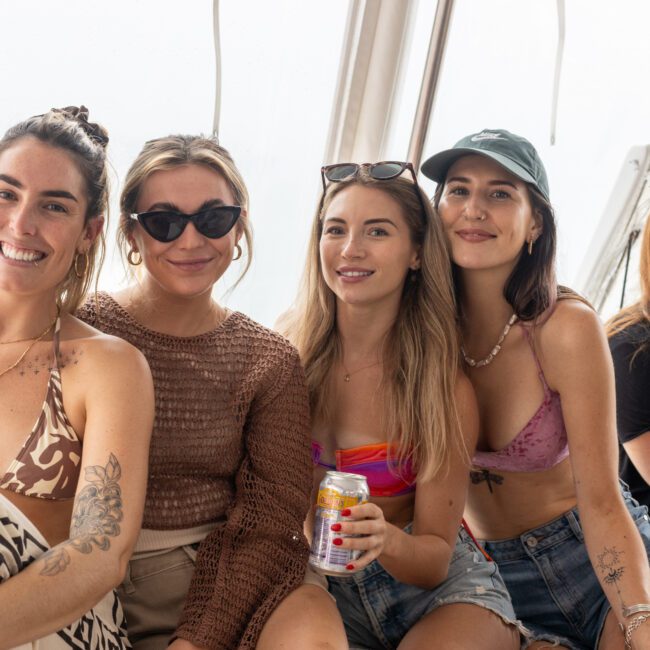 Five women sit together on a boat, smiling at the camera. Some are wearing bikinis, sunglasses, and casual clothing. One woman holds a can of drink. The bright background suggests it's daytime.