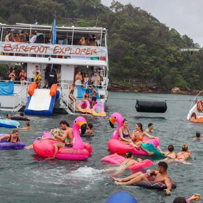 A lively scene of people enjoying a boat party on the water. Several individuals float on colorful inflatables, including flamingos and unicorns, near a large yacht named "Barefoot Explorer." The backdrop showcases lush, green hills under a bright blue sky.