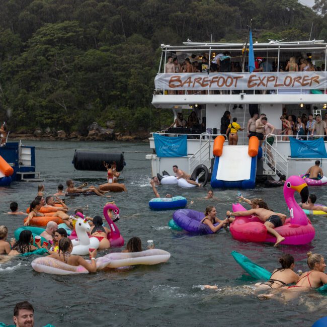 A lively scene of people enjoying a day on the water with a double-decker party boat named "Barefoot Explorer" in the background. The water is filled with individuals on colorful inflatables, including flamingos, unicorns, and donuts, all having fun kite surfing near the boat.