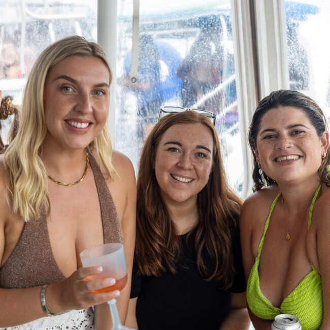 Three women smiling and posing for a photo on a boat. The woman on the left is holding a drink, the woman in the middle has red hair and is wearing a black top, and the woman on the right is in a green swimsuit holding a can. The background captures other passengers enjoying their time as well.