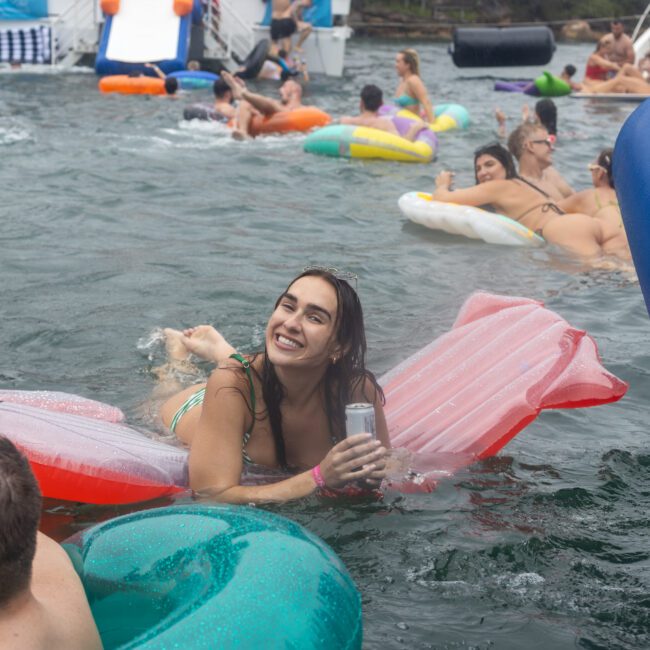 A woman is lounging on a pink float in a water body, smiling and holding a beverage can. Around her, people on various inflatable floats enjoy the water under the sunny sky. A boat is visible in the background. The atmosphere is lively and relaxed, suggestive of a fun summer social gathering.