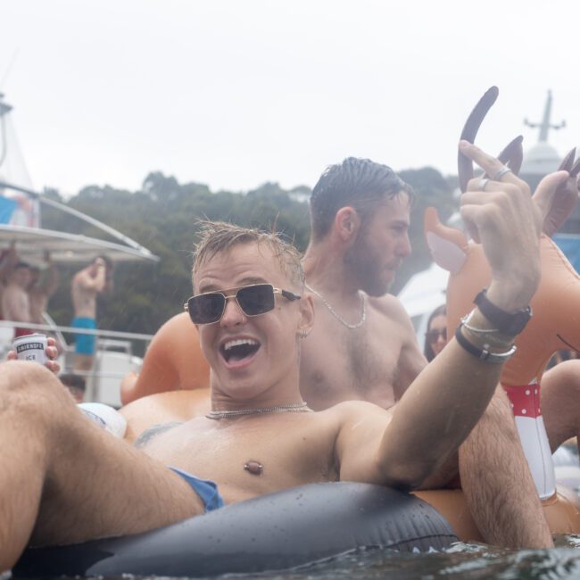 Three people enjoy a pool party on inflatable floaties; one enthusiastically poses with a peace sign, wearing sunglasses. The background features a boat with festive decorations and more partygoers in a cheerful atmosphere, complete with colorful fireworks lighting up the sky.