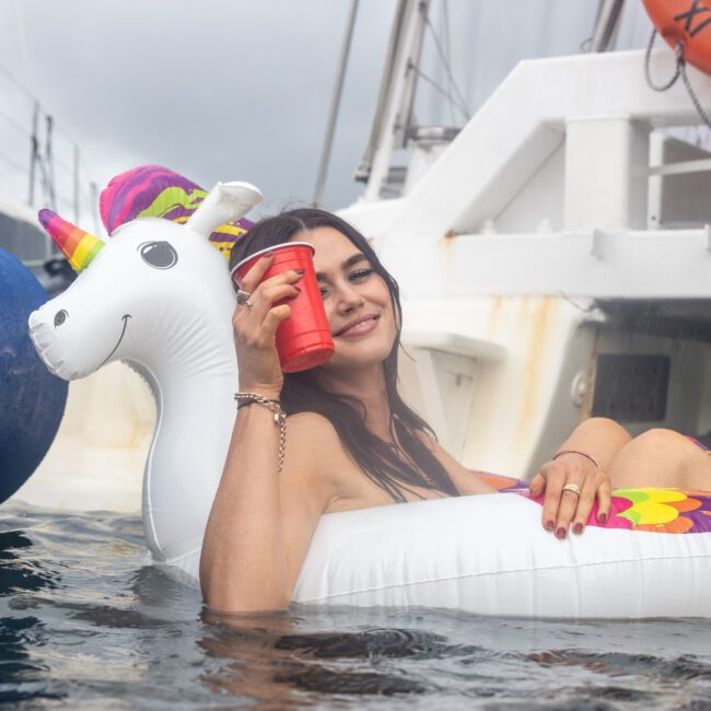 A woman relaxes in a pool while floating on an inflatable unicorn. She holds a red cup and smiles at the camera, with a boat and palm trees in the background. She appears to be enjoying a leisurely day in paradise.