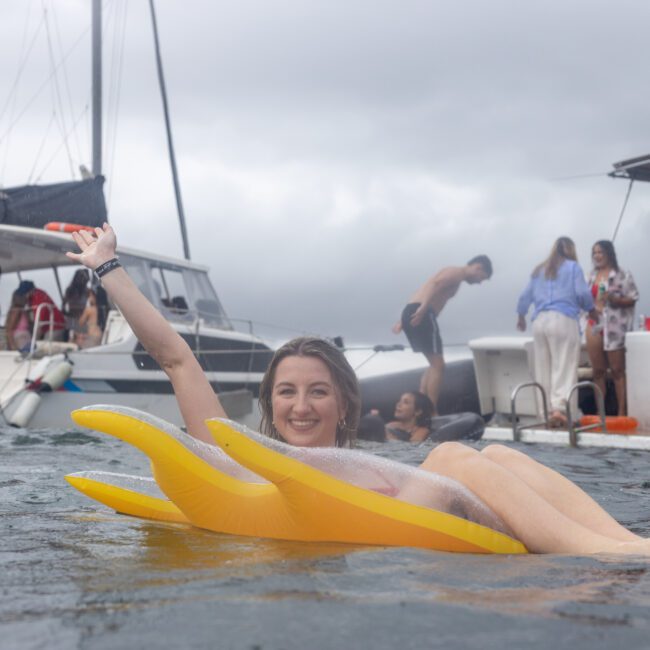 A woman floats on a yellow inflatable raft in the water, raising her arm and smiling. In the background, people are socializing on two docked boats under a cloudy sky. Solar panels can be seen on one of the boat's roofs.