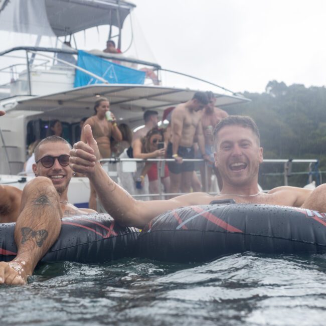 Two men are relaxing on inflatable tubes in the water, smiling and giving a thumbs-up. One man holds a refreshing drink. Behind them, people are gathered on a boat, some on the upper deck enjoying the festive atmosphere. Trees are visible in the background, completing the lively scene.