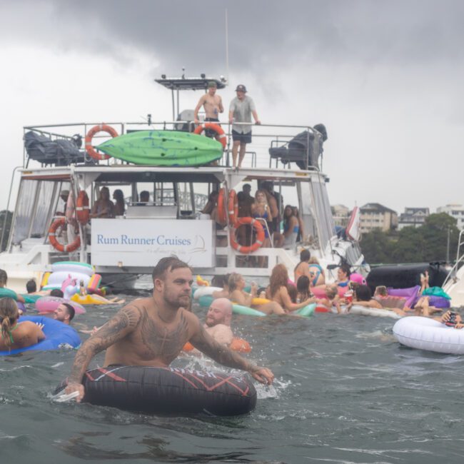 A group of people are enjoying a boat party on the water, with many floating on inflatable rings near a boat named "Rum Runner Cruises." The sky is overcast, and some people are on the deck while others splash around in the water, socializing and having fun. Laughter fills the air as everyone delights in the lively atmosphere.