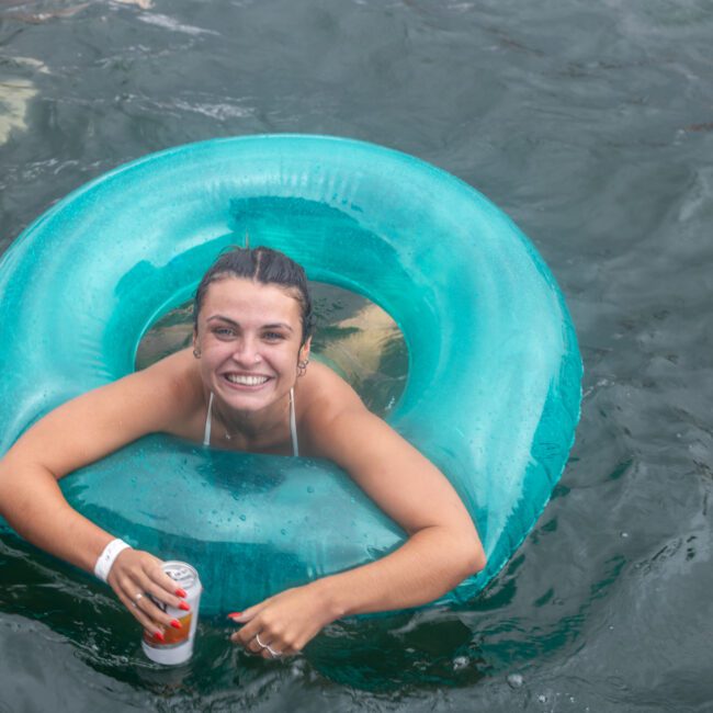 A smiling person with dark hair is partially submerged in water, floating inside a turquoise inner tube. They are holding a can in their right hand and wearing a white strapless top. The wavy water around them adds to the vibrant scene, with other indistinct figures enjoying the background.