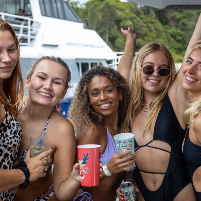 A group of five smiling women in swimsuits pose for a photo on a yacht. They are holding drinks, enjoying a day on the water, with a large white boat and lush greenery visible in the background.