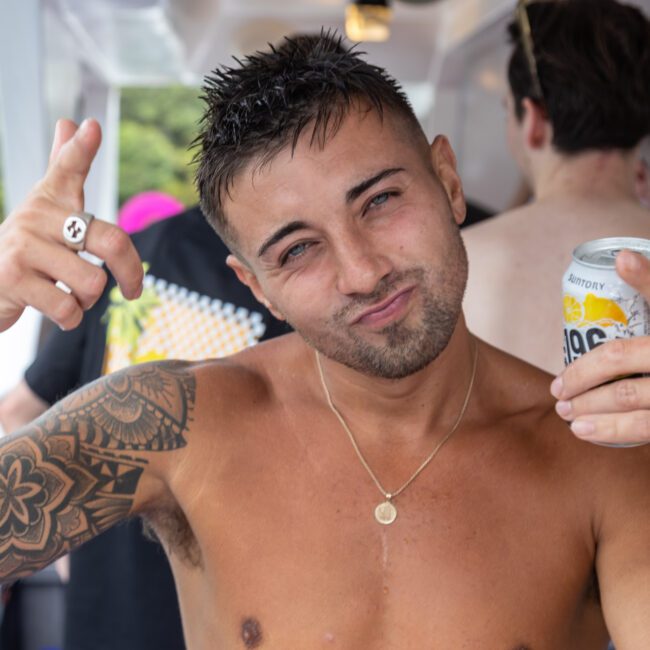 A shirtless man with tattoos and a necklace poses for the camera, holding a refreshing drink in his right hand and pointing with his left. Other shirtless people are blurred in the background. The atmosphere seems casual and social, giving off relaxed vibes.