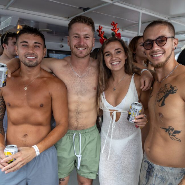 Four people smile and pose for a group photo at a boat party. Two shirtless men on the left and right display tattoos, while a man in green shorts and a woman in a white dress with red antlers stand in the middle. Festive lights illuminate the background with other partygoers enjoying themselves.