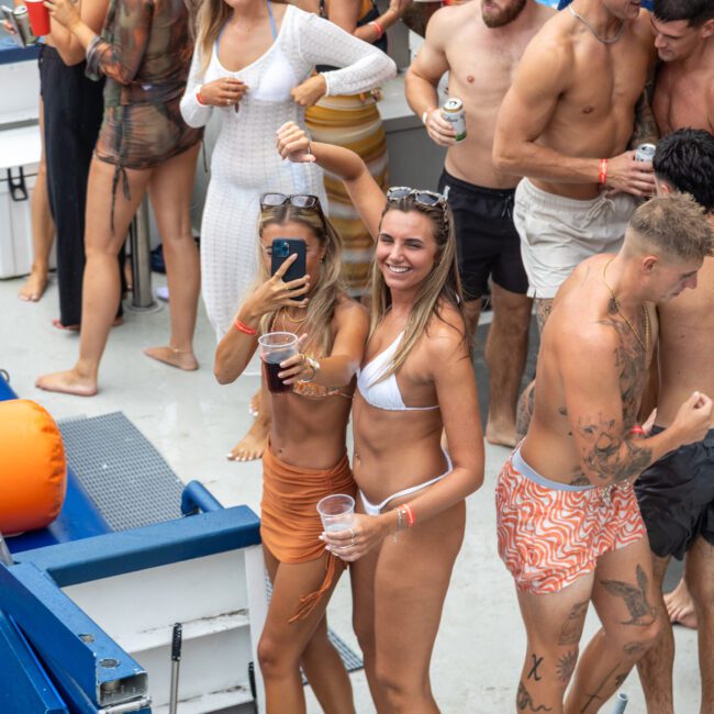 A lively group of young people enjoying a party on a boat. Some are in swimwear, holding drinks, and dancing. There are inflatable toys, deck seating, and vibrant decorations visible. The atmosphere is energetic and festive.