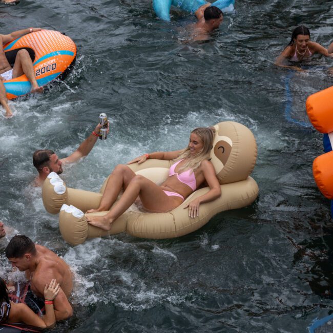 A group of people enjoying a pool party with various inflatables. A woman in a pink bikini lounges on a large monkey-shaped float while others swim and socialize around her. A man holding a drink floats nearby, next to an orange and blue inflatable slide in the corner.